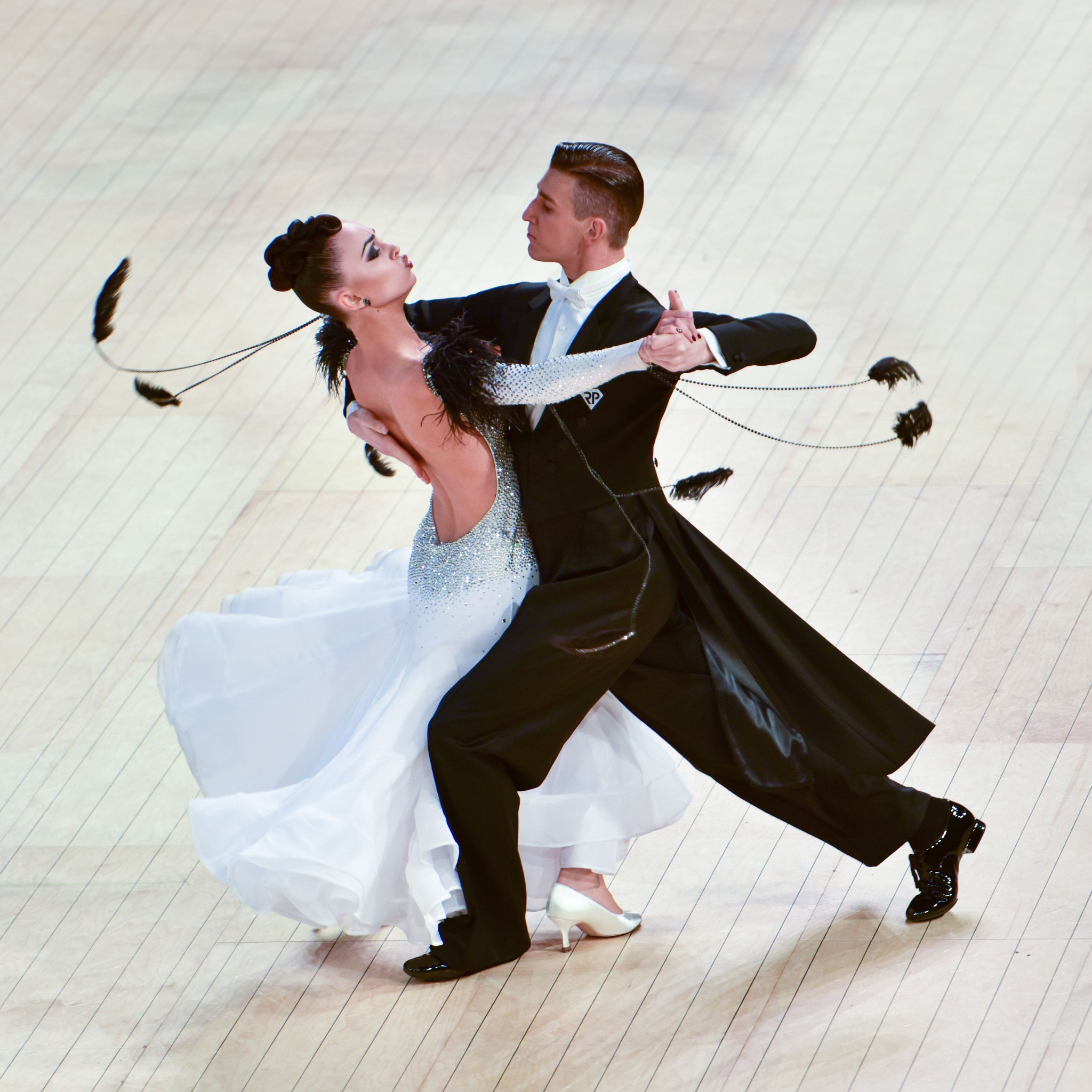 Para tancerzy w strojach galowych prezentuje taniec towarzyski na parkiecie z jasnego drewna.