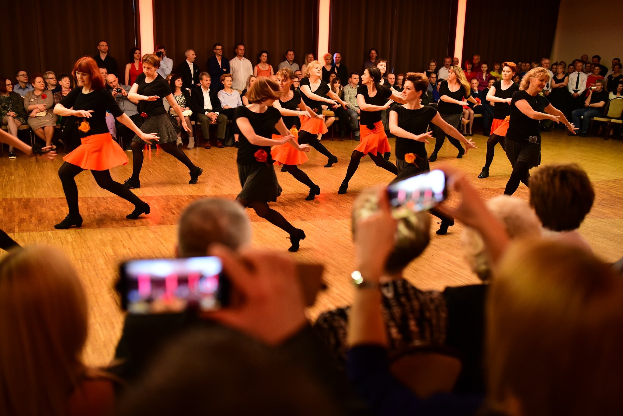 Grupa kobiet w czarnych bluzkach i pomarańczowych spódnicach tańczy na parkiecie, widzowie obserwują i filmują występ smartfonami.