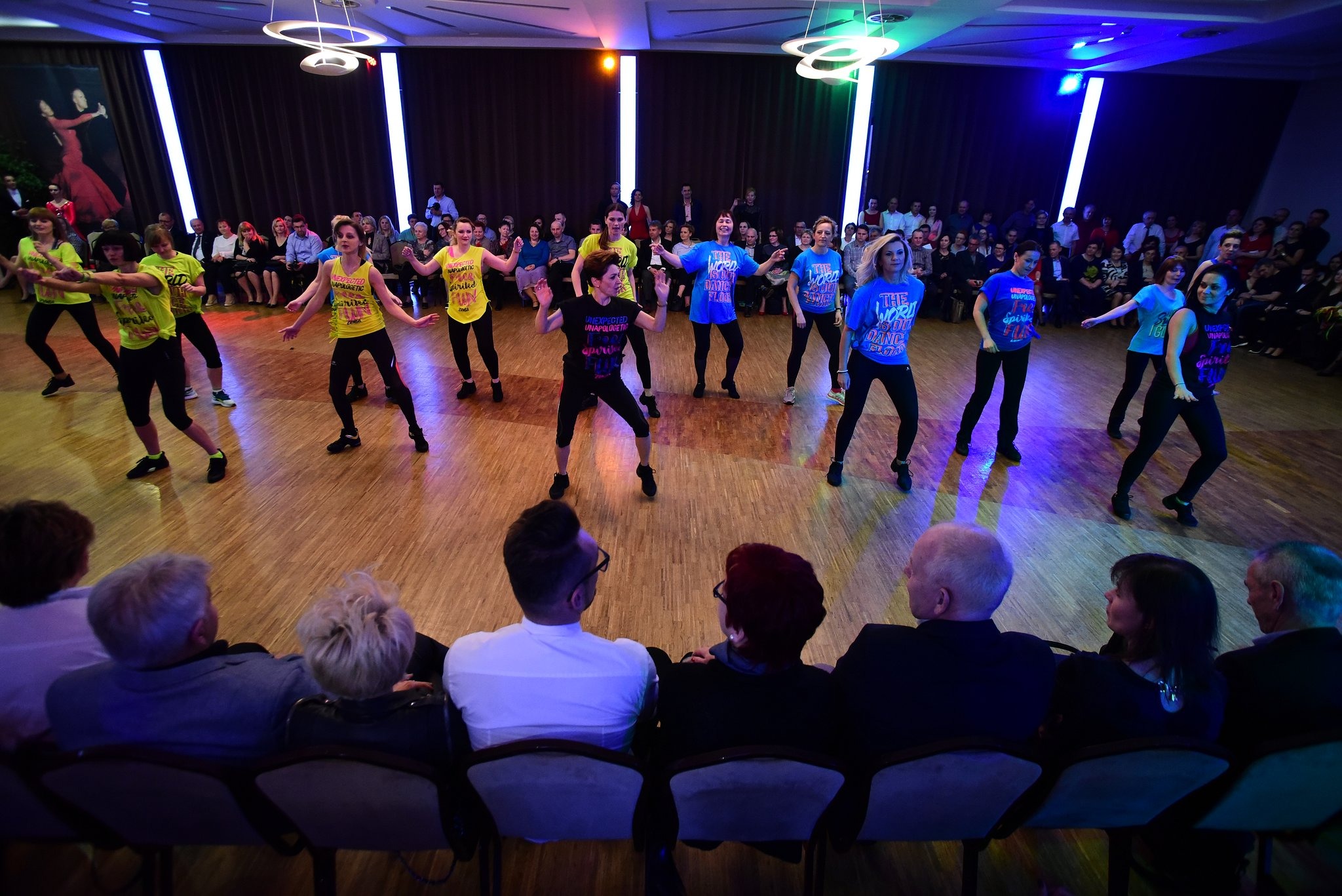 Grupa kobiet w kolorowych koszulkach tańczy na parkiecie przed publicznością, oświetlenie sceniczne tworzy dynamiczne efekty świetlne.
