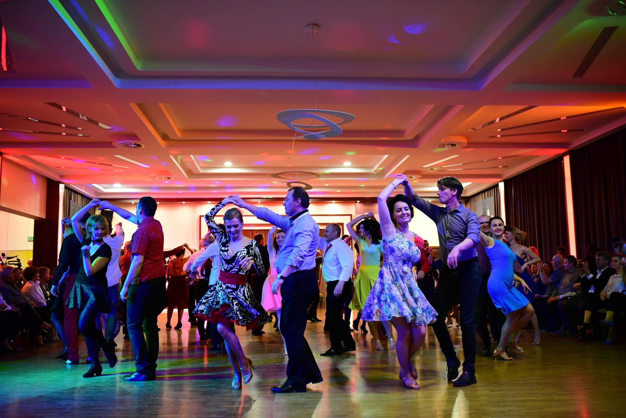 Grupa osób tańczących w sali balowej oświetlonej kolorowymi światłami, z parkietem odbijającym światło i publicznością siedzącą w tle.