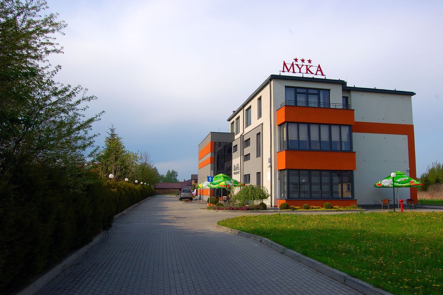 Widok zewnętrzny nowoczesnego, trzypiętrowego budynku z pomarańczowymi akcentami, zielonymi parasolami i brukowanym podjazdem, nazwa 'MYKA' z trzema gwiazdkami umieszczona na dachu.
