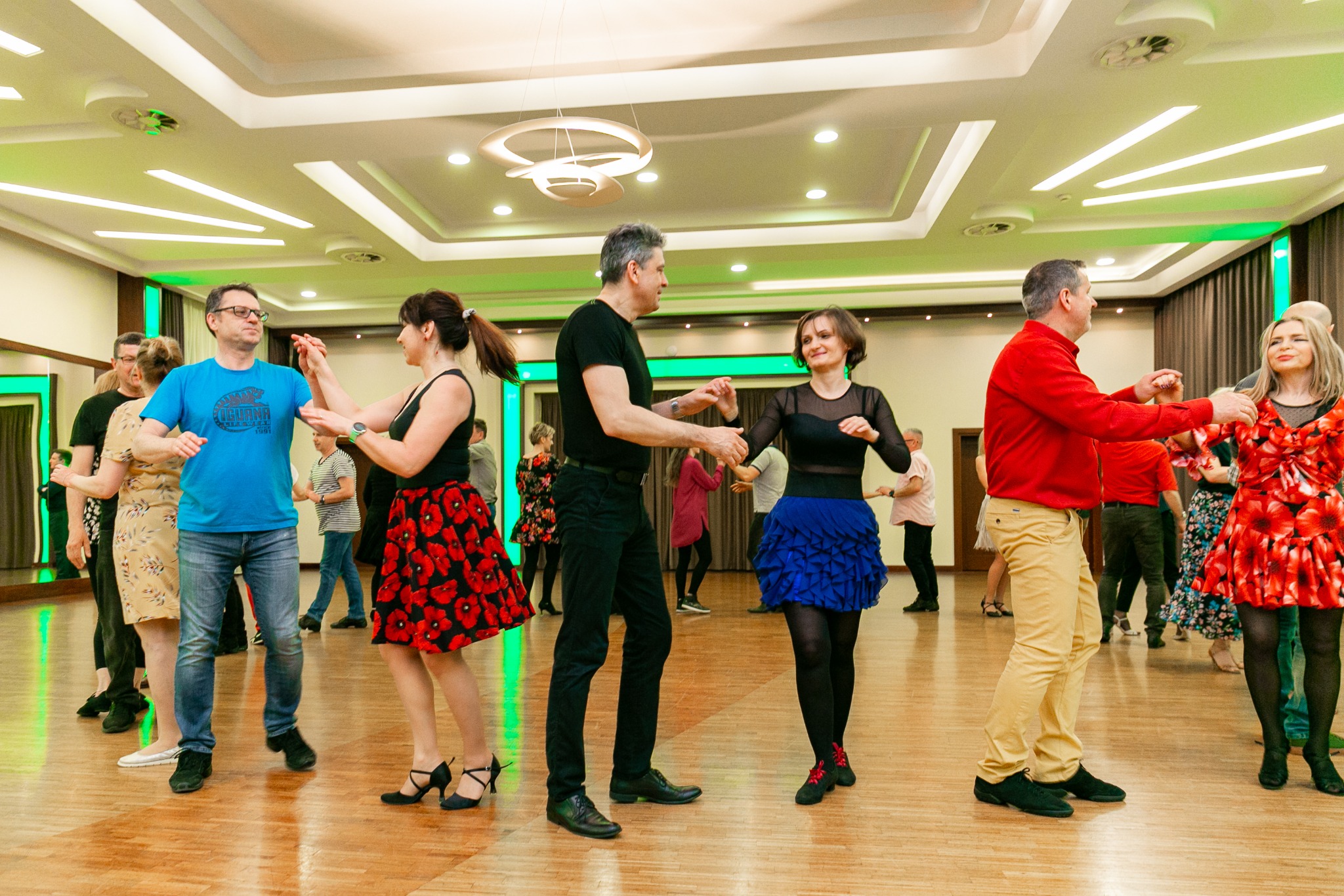 Grupa osób w różnym wieku tańczy w sali z parkietem i oświetleniem LED, kobiety w kolorowych sukienkach, mężczyźni w swobodnych strojach.