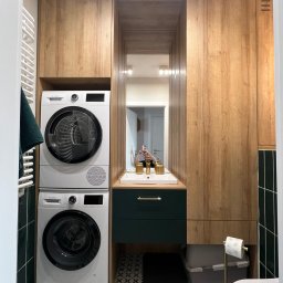 Meble Owczarek - Nowoczesna łazienka z drewnianą zabudową meblową, pralką i suszarką w kolumnie, zieloną szafką pod umywalką z złotą armaturą, widoczne odbicie osoby robiącej zdjęcie w lustrze.