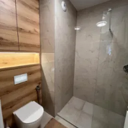 Nowoczesna łazienka z drewnianą zabudową, podświetlaną wnęką, podwieszaną toaletą z bidetką i kabiną prysznicową z marmurowymi płytkami.