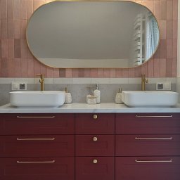Meble Owczarek - Nowoczesna łazienka z burgundową szafką pod umywalką, dwiema białymi umywalkami nablatowymi, złotymi bateriami i podłużnym lustrem w złotej ramie, oświetlonym kulistymi lampami na tle różowych płytek.
