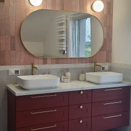 Meble Owczarek - Nowoczesna łazienka z podwójną umywalką nablatową, bordowymi szafkami z złotymi uchwytami, różowymi płytkami na ścianie i owalnym lustrem z podświetleniem.