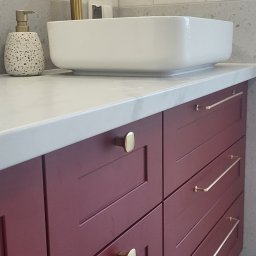 Meble Owczarek - Bordowa szafka pod umywalkę z białym blatem i złotą armaturą, widoczny dozownik do mydła w kropki i okrągłe lustro z podświetleniem.