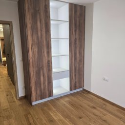 Meble Owczarek - Szafa wnękowa z frontami w kolorze ciemnego drewna, z oświetleniem LED wewnątrz, na tle parkietu i białych ścian.