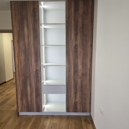 Meble Owczarek - Szafa wnękowa z oświetleniem LED, brązowe fronty imitujące drewno, białe wnętrze z półkami i szufladą, widok z przodu na tle jasnej ściany i drewnianej podłogi.