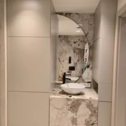 Nowoczesna łazienka z szafkami na wymiar w neutralnych barwach, marmurowym blatem i okrągłym lustrem. Umywalka nablatowa i czarna bateria dodają elegancji.