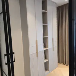 Jasna szafa wnękowa z ryflowanymi frontami i otwartymi półkami, widoczna z perspektywy wejścia, z zasłoną w tle i parkietem na podłodze.