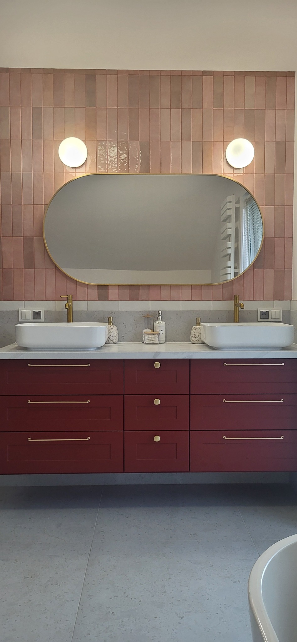 Nowoczesna łazienka z burgundową szafką pod umywalką, dwiema białymi umywalkami nablatowymi, złotymi bateriami i podłużnym lustrem w złotej ramie, oświetlonym kulistymi lampami na tle różowych płytek.