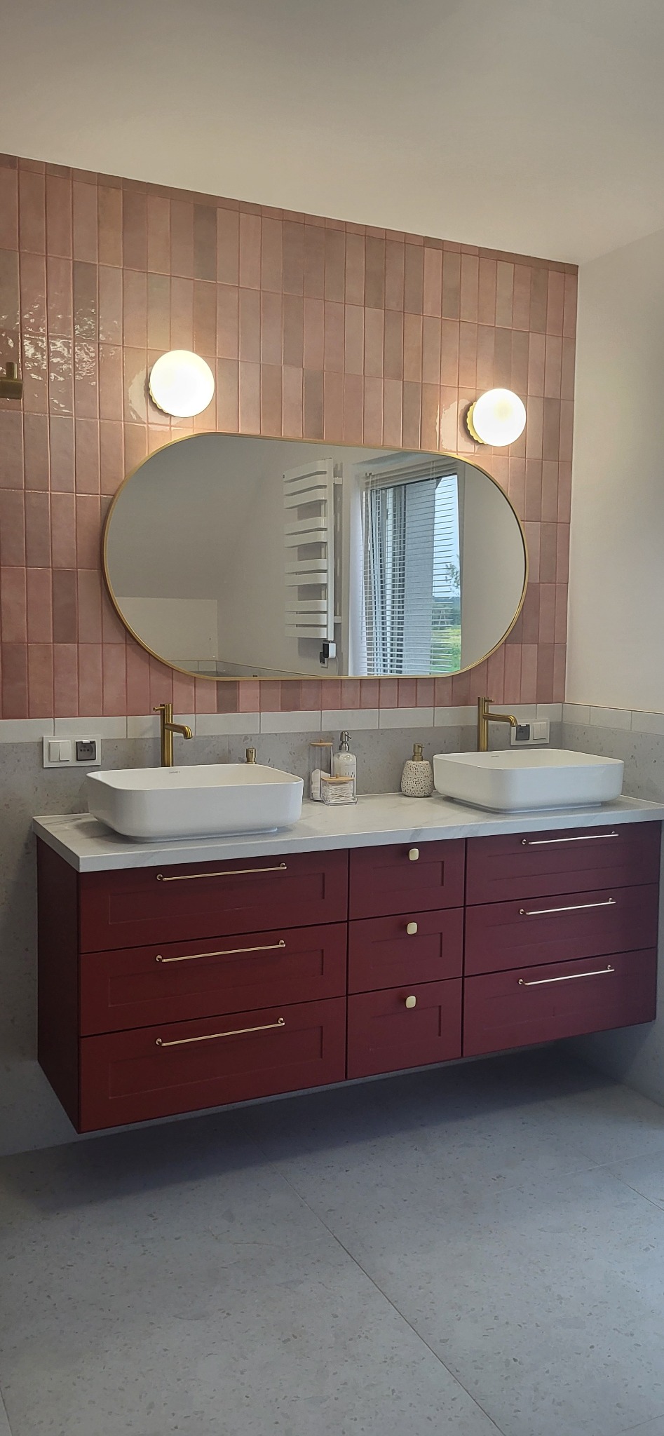 Nowoczesna łazienka z podwójną umywalką nablatową, bordowymi szafkami z złotymi uchwytami, różowymi płytkami na ścianie i owalnym lustrem z podświetleniem.