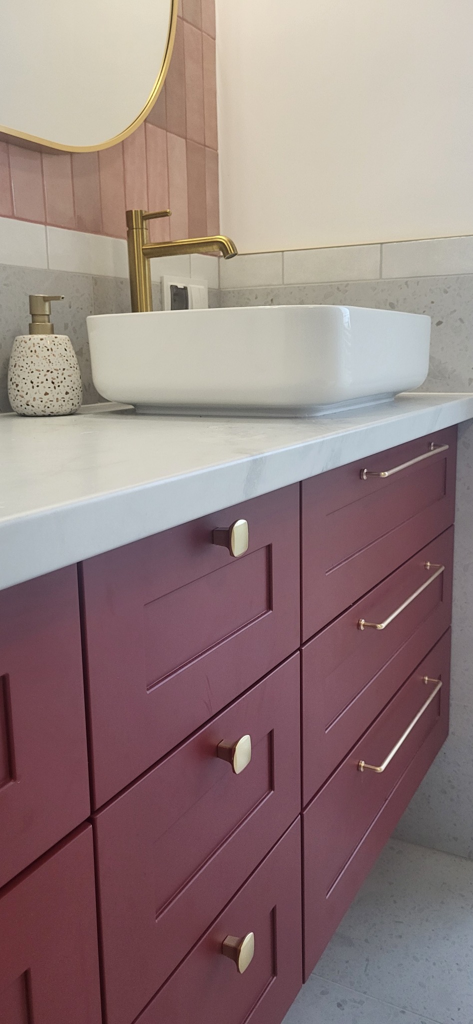 Bordowa szafka pod umywalkę z białym blatem i złotą armaturą, widoczny dozownik do mydła w kropki i okrągłe lustro z podświetleniem.