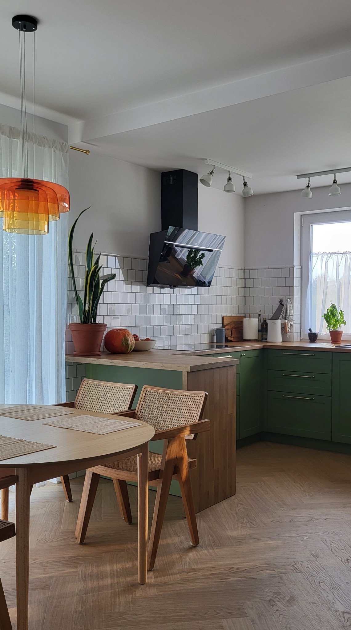 Jasna kuchnia z zielonymi szafkami, drewnianym blatem i wyspą, okrągły stół z krzesłami z rattanowym siedziskiem, lampa w odcieniach pomarańczy i żółci.