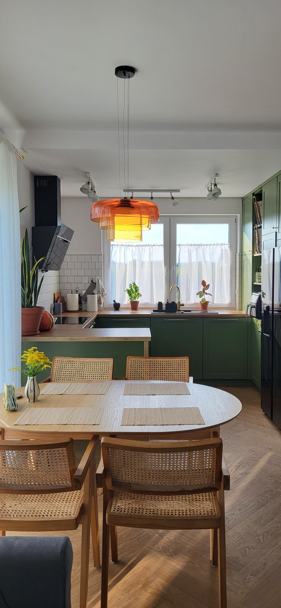 Wnętrze kuchni z zielonymi szafkami, drewnianym blatem, okrągłym stołem i krzesłami z rattanowym siedziskiem, nad stołem pomarańczowa lampa.