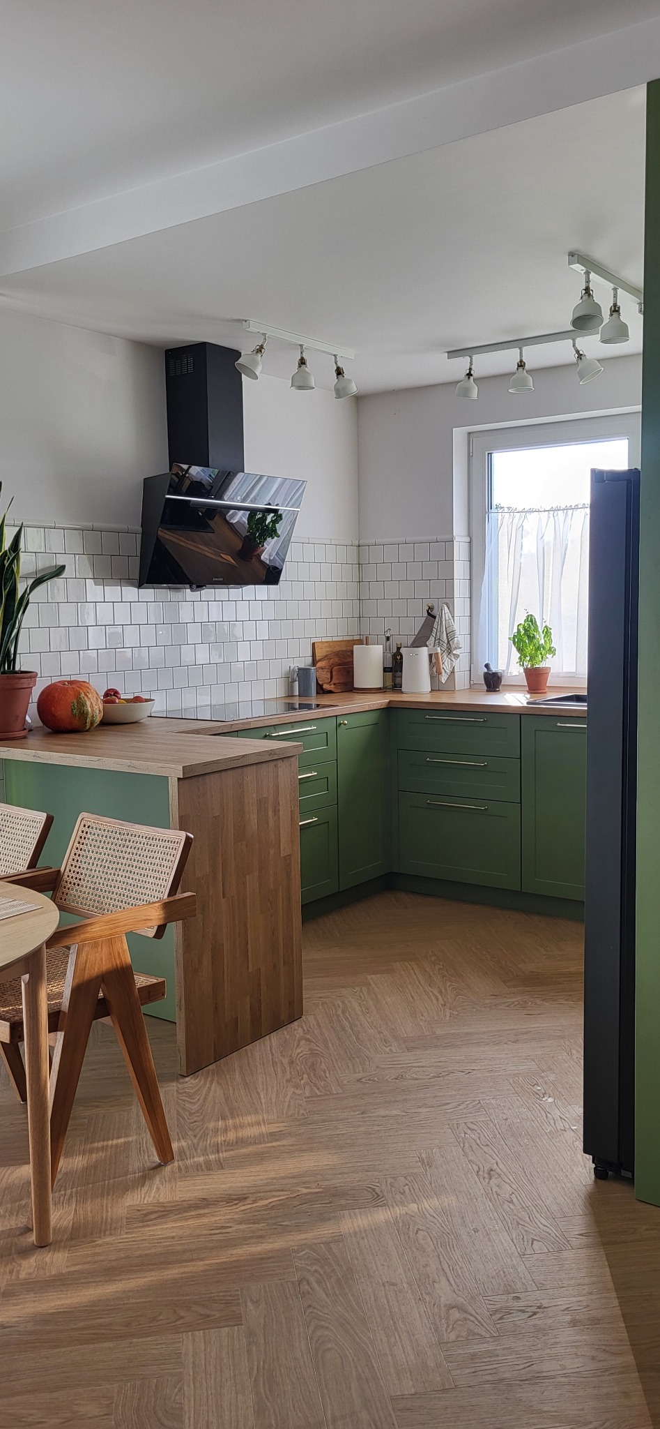 Zielona kuchnia z drewnianym blatem, białymi kaflami i czarnym okapem, oświetlona punktowymi lampami, z drewnianym stołem i krzesłami w rogu, na podłodze parkiet ułożony w jodełkę.