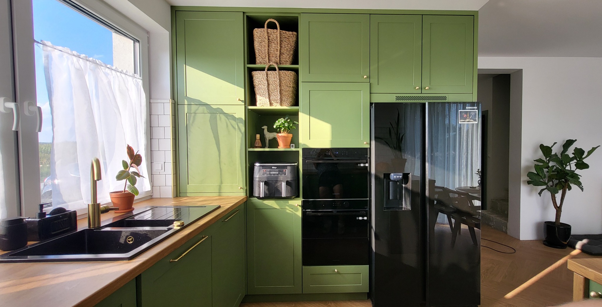 Nowoczesna kuchnia w odcieniach zieleni z drewnianym blatem, złotą armaturą i czarnym zlewem, uzupełniona o czarną lodówkę side-by-side i wbudowany piekarnik, z wiklinowymi koszami i roślinami...