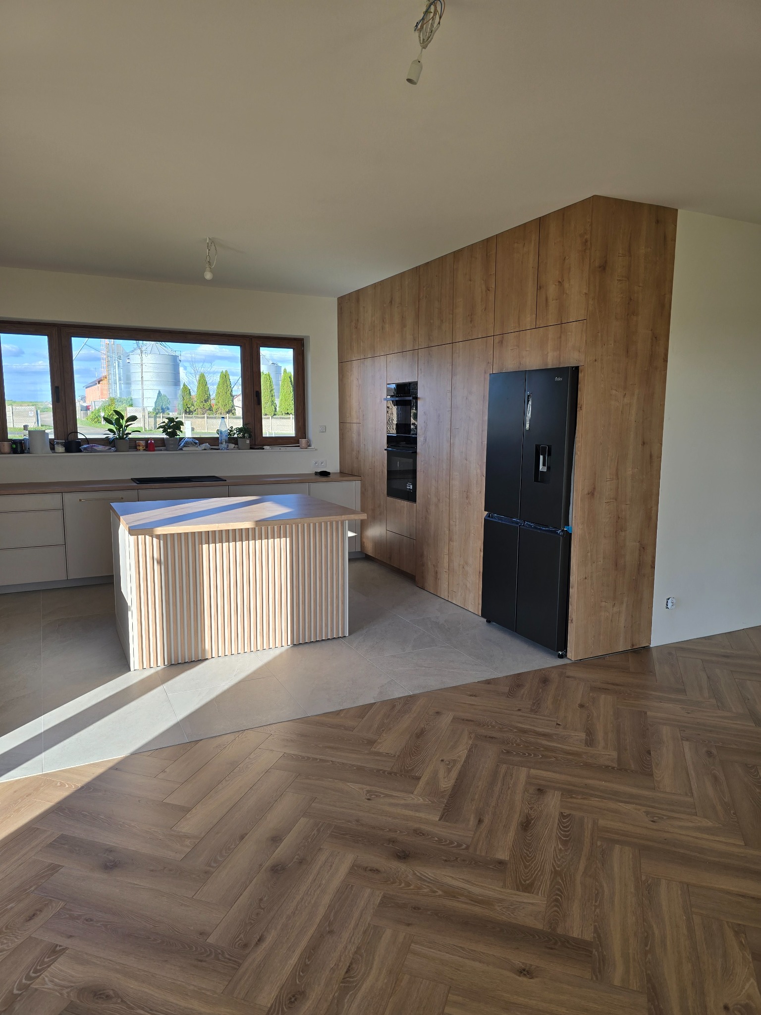 Nowoczesna kuchnia z wyspą i zabudową w odcieniach drewna, czarna lodówka. Parkiet w jodełkę, duże okno z widokiem na silosy. Jasne, przestronne wnętrze.
