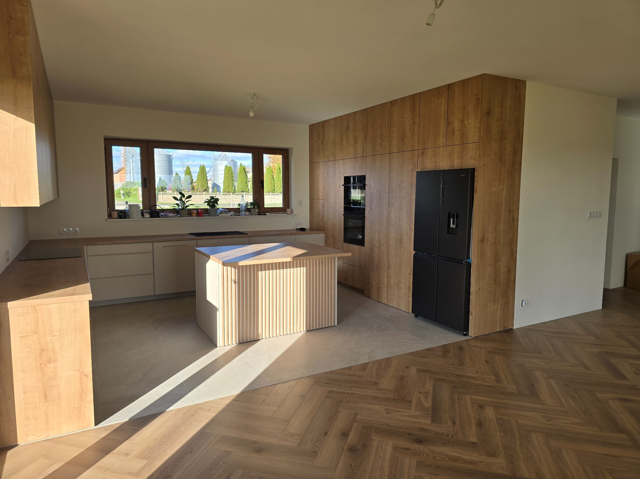 Jasna kuchnia z drewnianymi frontami, wyspą i czarną lodówką. Duże okno z widokiem na silosy. Parkiet w jodełkę. Nowoczesne i przestronne wnętrze.
