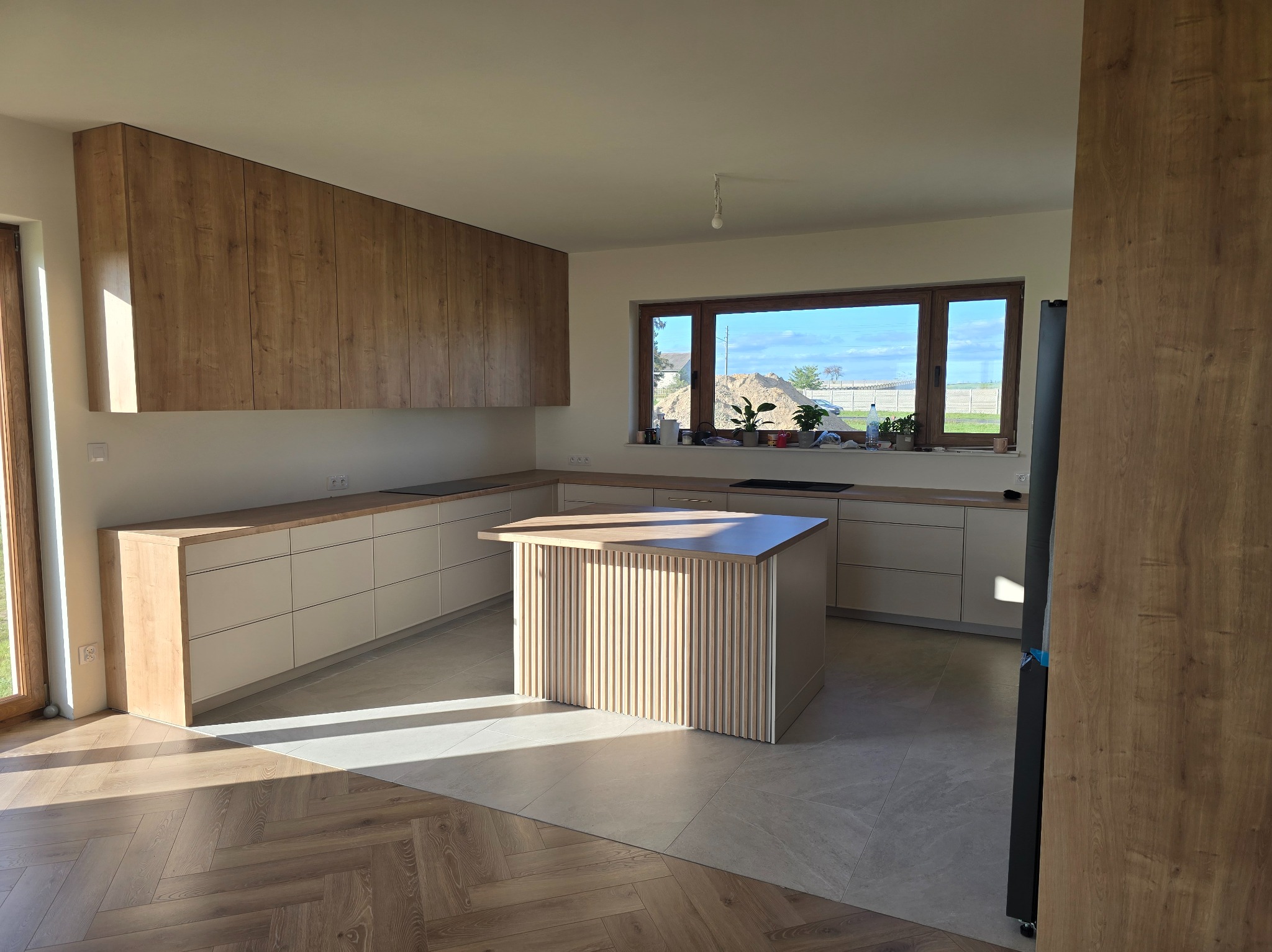 Jasna kuchnia z drewnianymi szafkami wiszącymi i wyspą z lamelami, okno z widokiem na zewnątrz. Na podłodze parkiet i płytki. Wnętrze doświetlone naturalnym światłem.