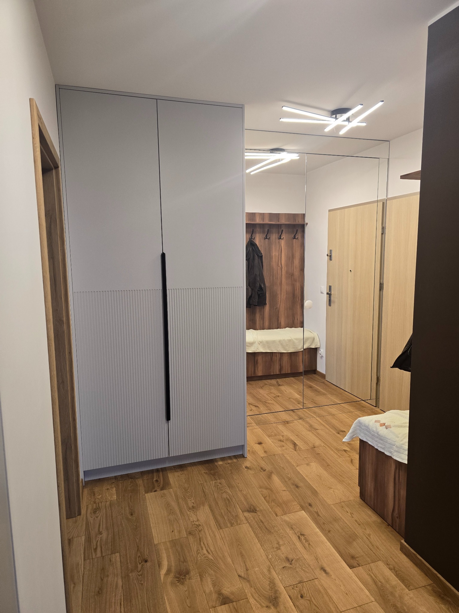 Szafa wnękowa z szarymi frontami i pionowymi żłobieniami, czarne uchwyty, lustro w przedpokoju, drewniana podłoga. Widok na wieszak i siedzisko w odbiciu lustra.