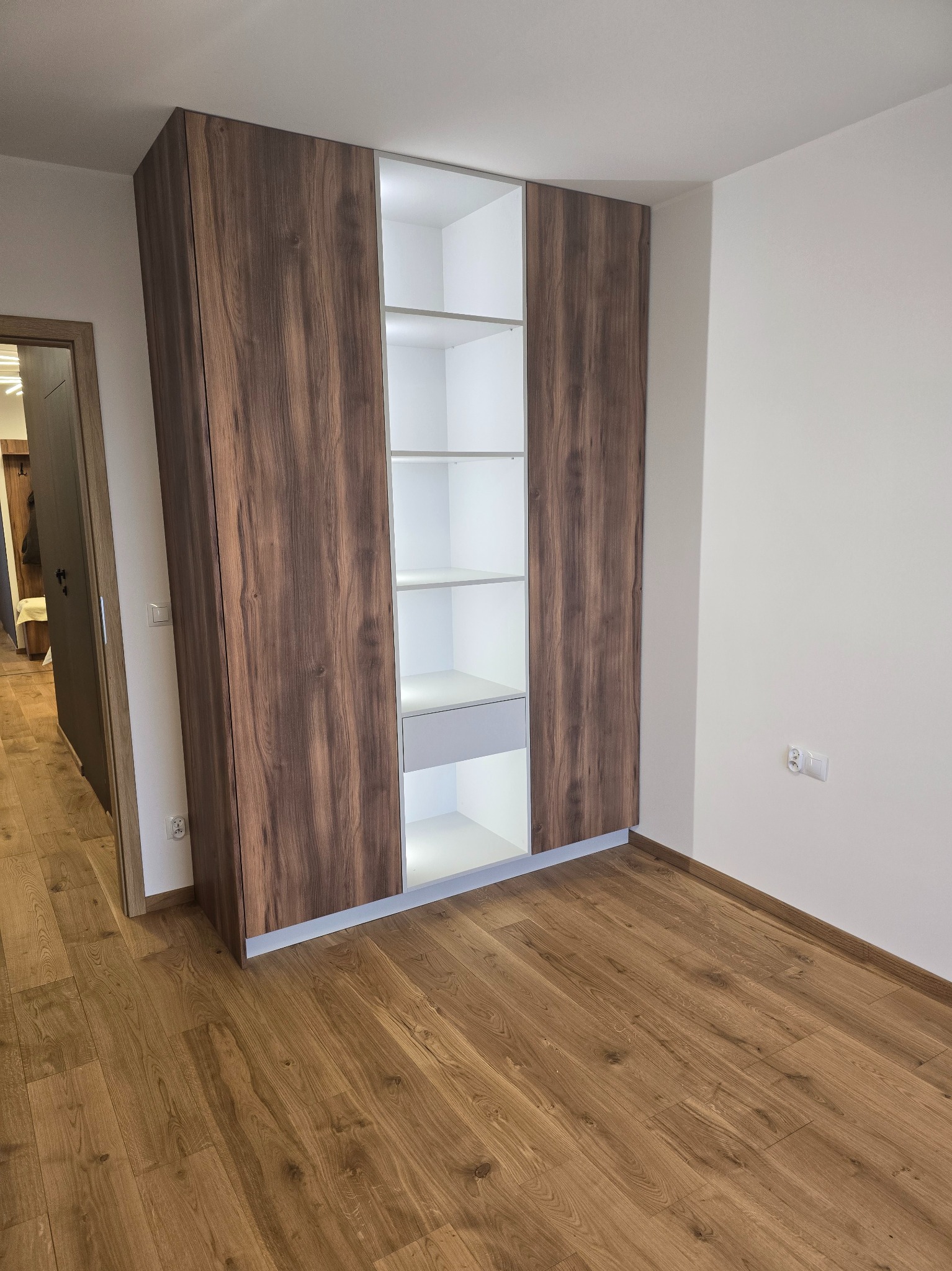 Szafa wnękowa z frontami w kolorze ciemnego drewna, z oświetleniem LED wewnątrz, na tle parkietu i białych ścian.