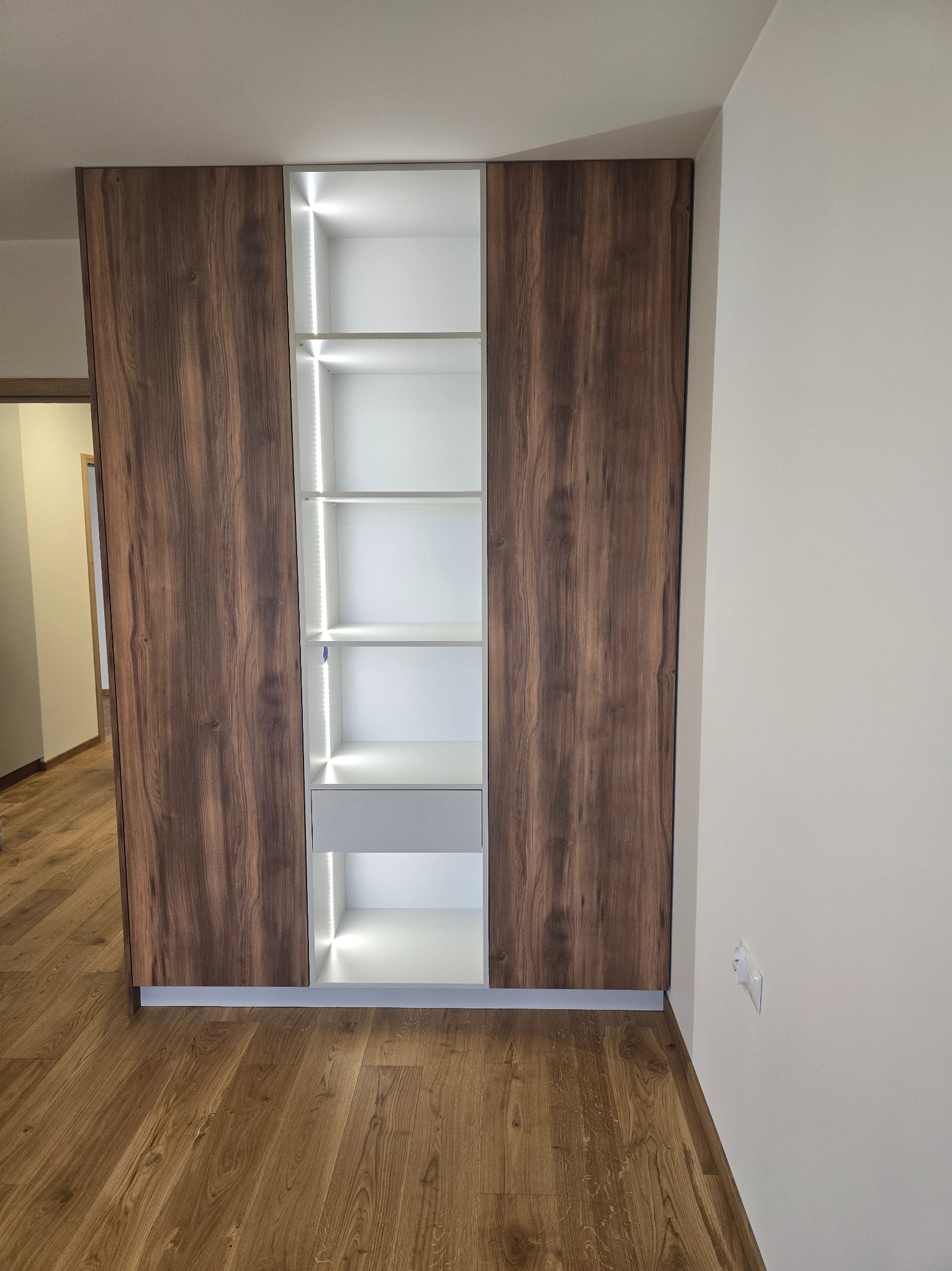 Szafa wnękowa z oświetleniem LED, brązowe fronty imitujące drewno, białe wnętrze z półkami i szufladą, widok z przodu na tle jasnej ściany i drewnianej podłogi.