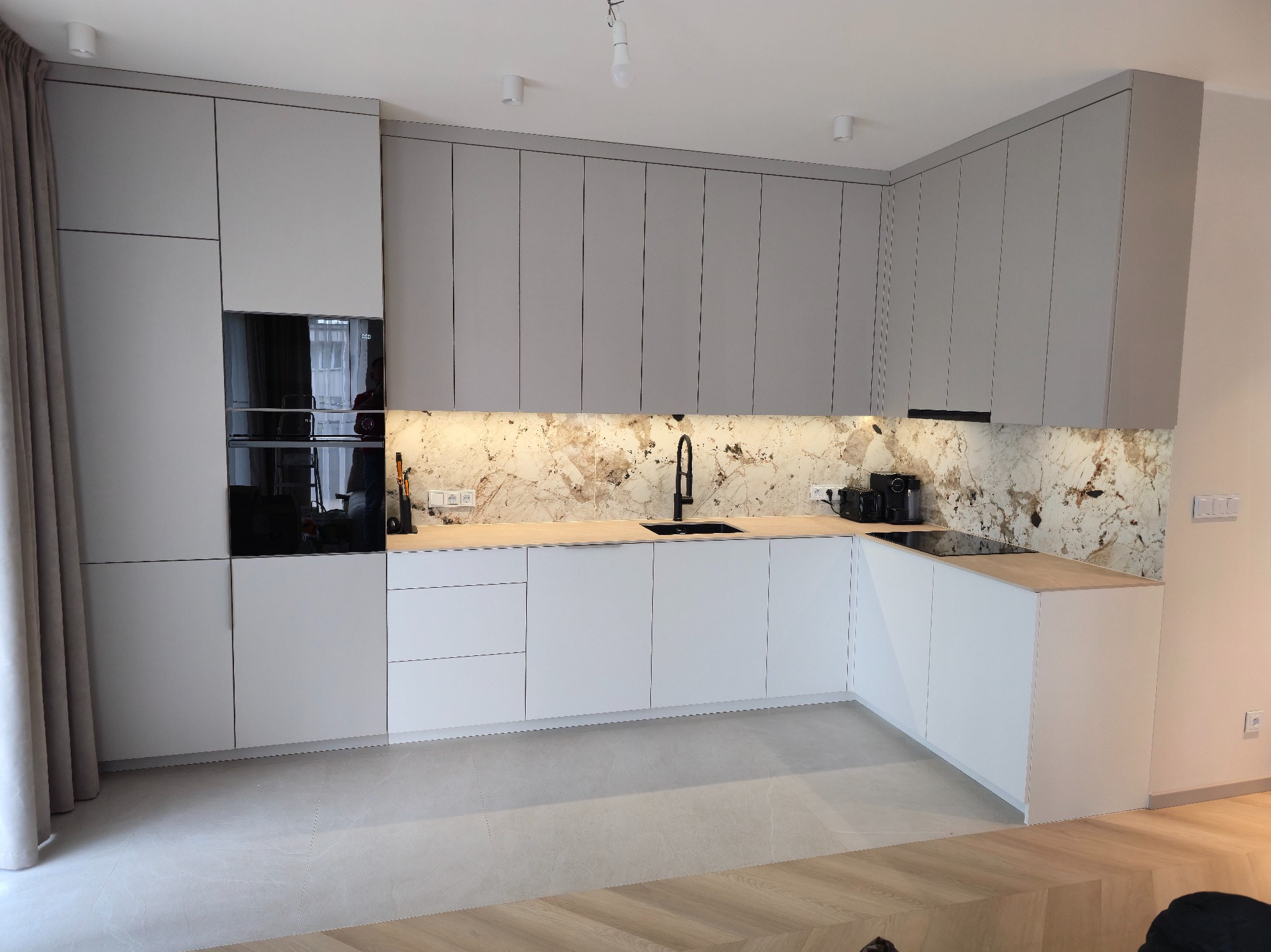 Nowoczesna kuchnia z szarymi szafkami górnymi i białymi dolnymi, marmurowym blatem i czarną armaturą. Widoczne sprzęty AGD i oświetlenie podszafkowe.