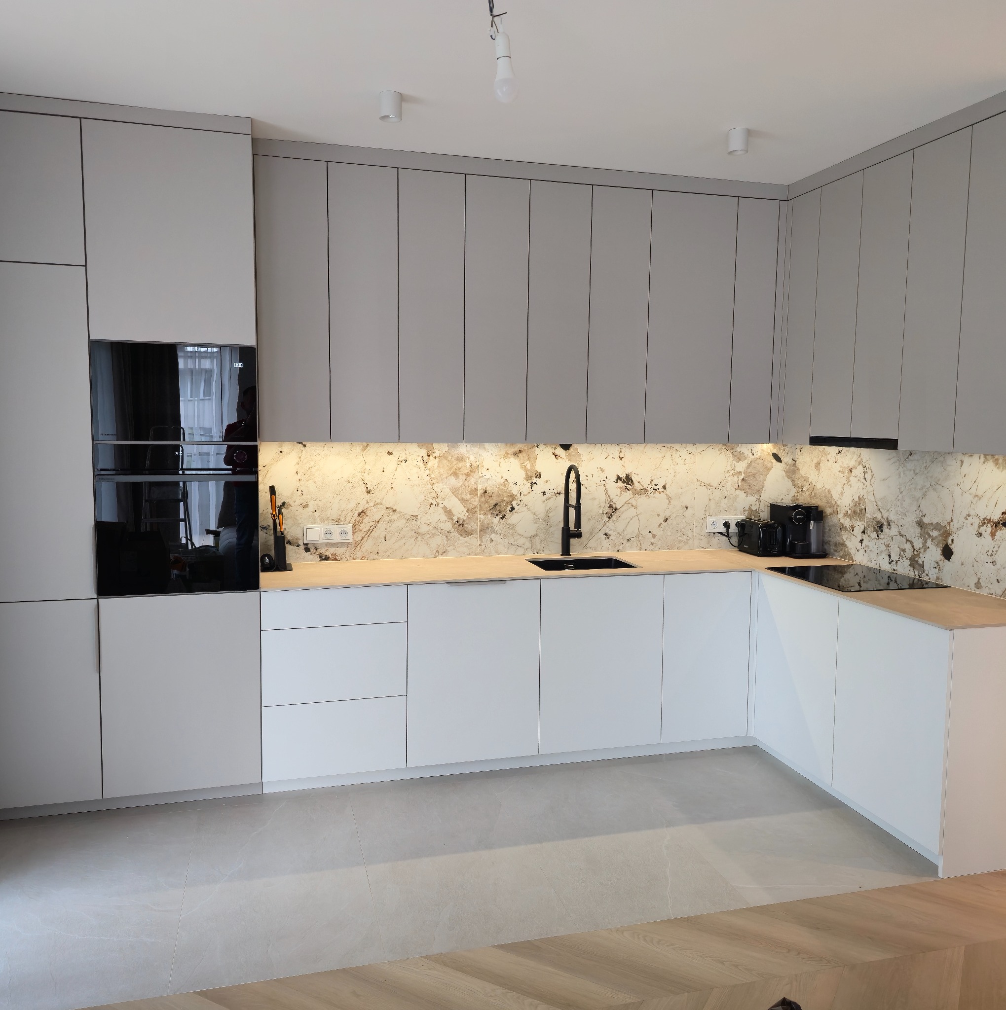 Nowoczesna kuchnia z szarymi szafkami górnymi i białymi dolnymi. Marmurowy backsplash podświetlony. Czarny zlew i sprzęt AGD. Parkiet w jodełkę.