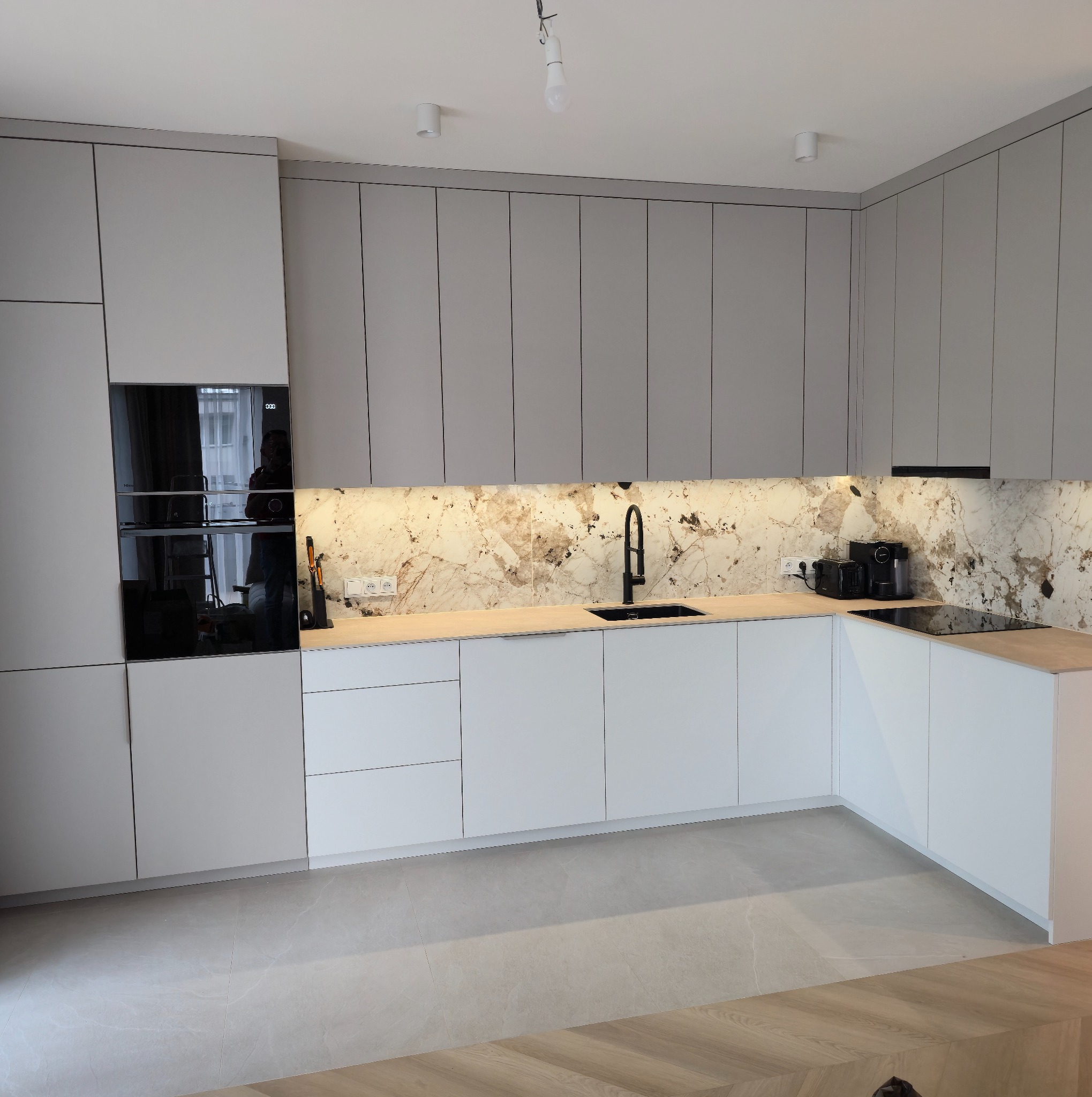 Nowoczesna kuchnia w odcieniach szarości i bieli, z marmurowym blatem i czarną armaturą. Widoczne sprzęty AGD i oświetlenie podszafkowe.