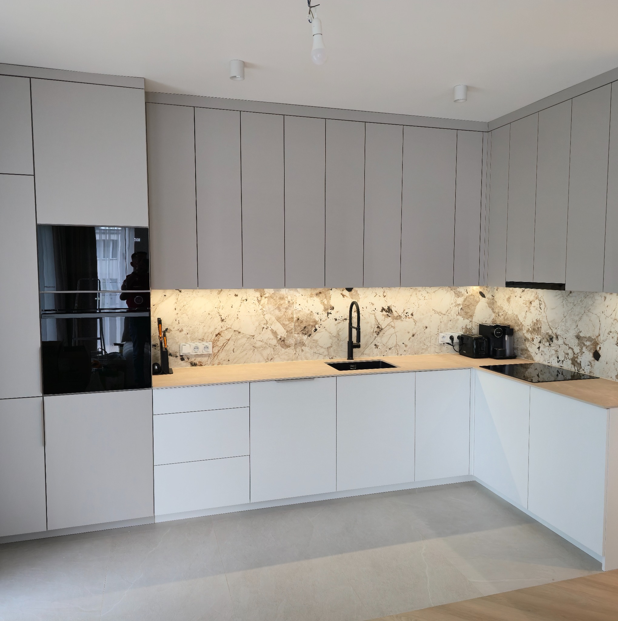 Nowoczesna kuchnia na wymiar z szarymi górnymi szafkami i białymi dolnymi. Marmurowy backsplash, czarna armatura i sprzęt AGD. Minimalistyczny design.