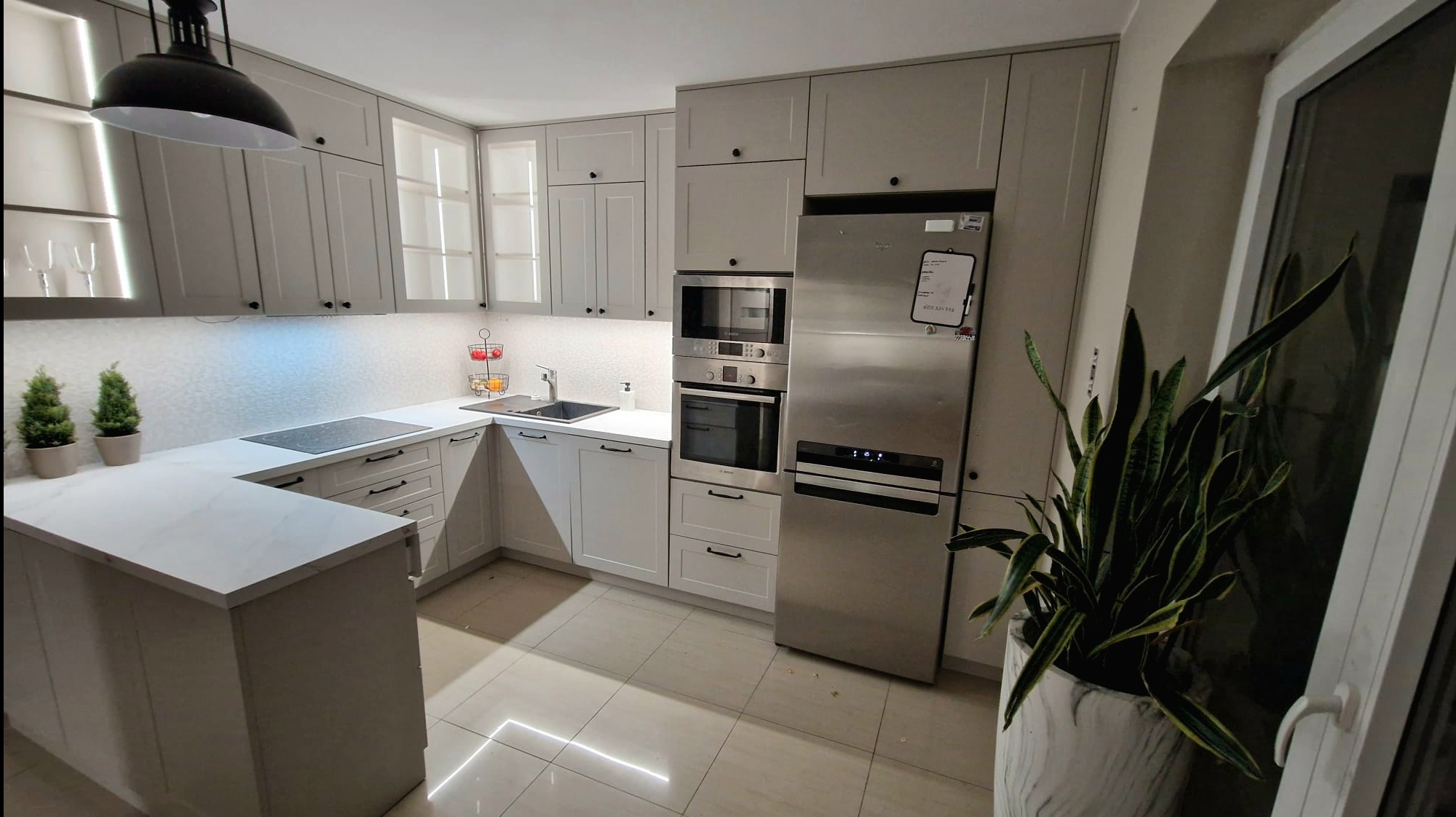 Jasna kuchnia z szarymi frontami, czarnymi uchwytami i sprzętem AGD w zabudowie. Uwagę przyciąga lampa nad blatem i roślina doniczkowa przy oknie.