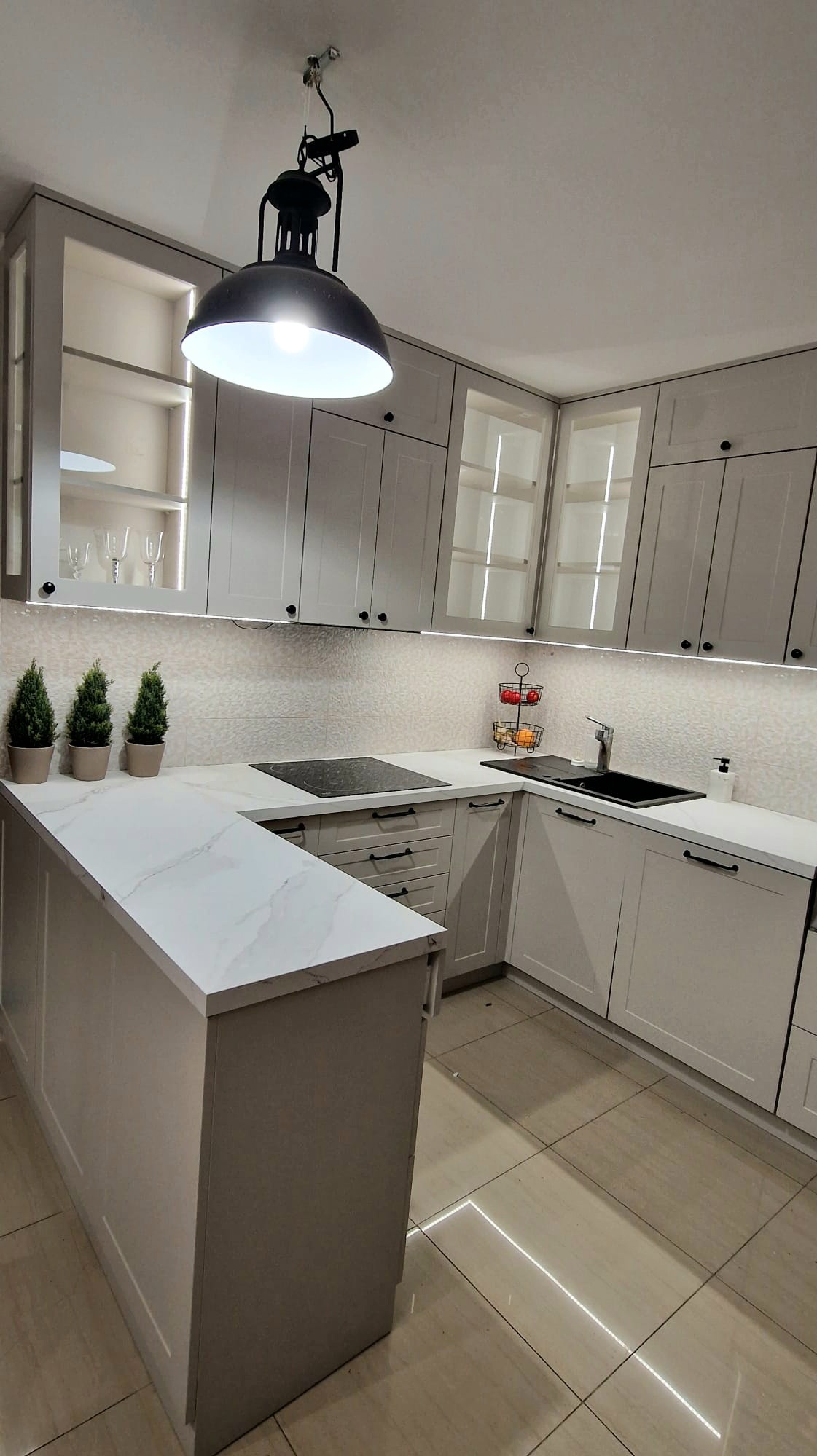 Jasna kuchnia z wyspą w Woli Jedlińskiej. Szafki z podświetleniem LED, czarny zlew i indukcja. Na blacie dekoracyjne drzewka i owoce.