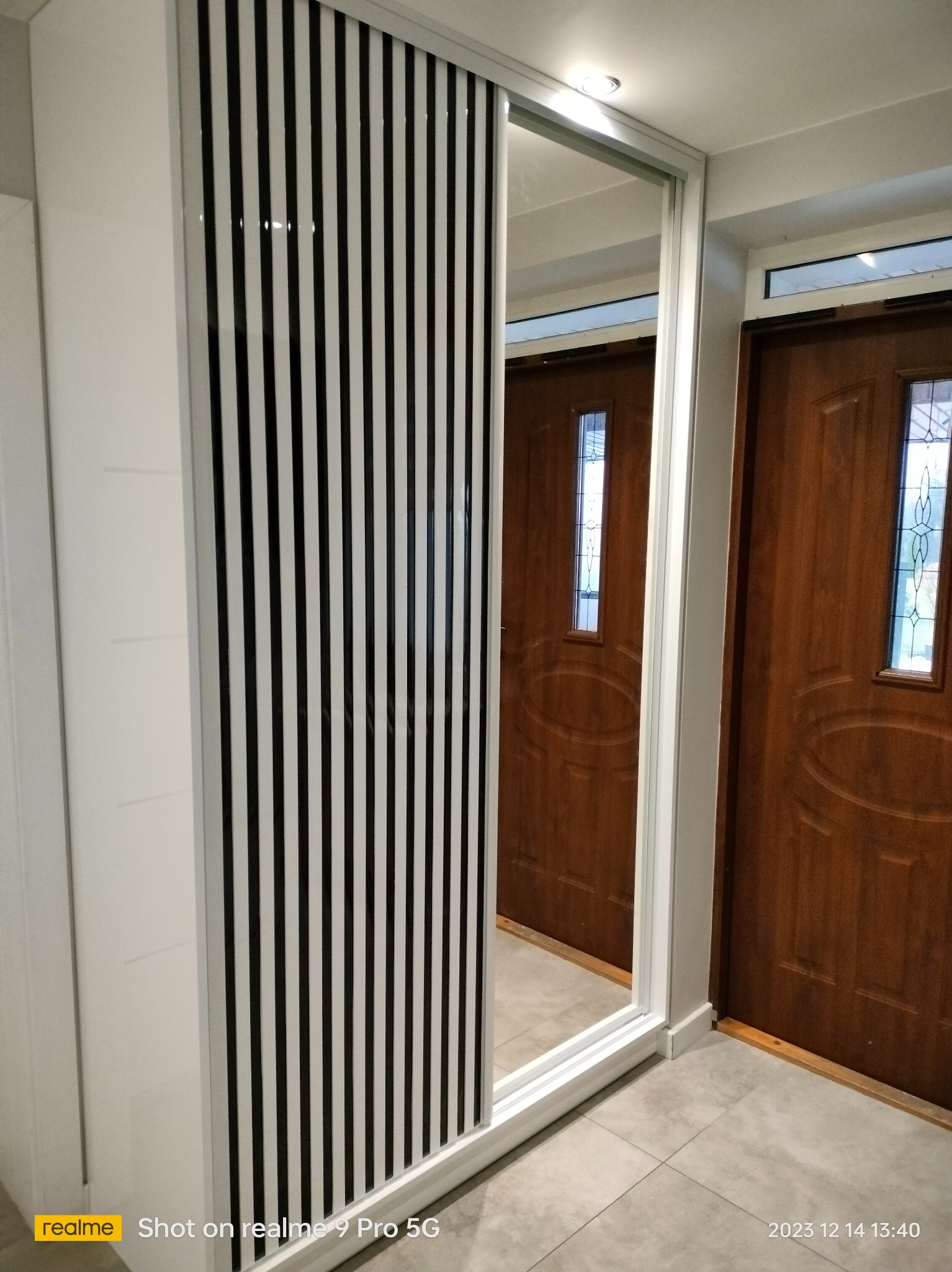 Biała szafa wnękowa z lustrem i frontem w czarno-białe pasy, w przedpokoju z brązowymi drzwiami. Minimalistyczny design, nowoczesne wykończenie.