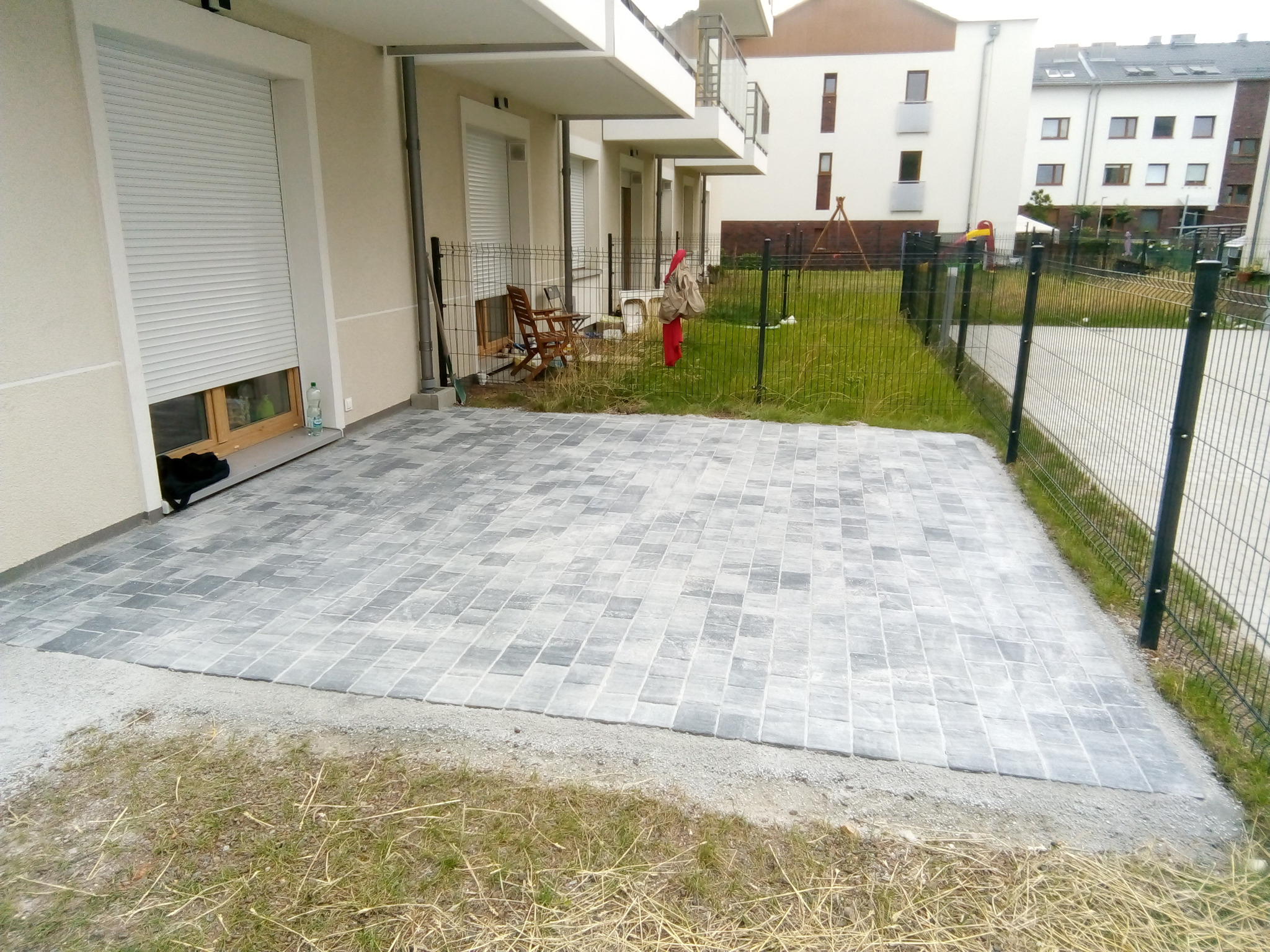 Ułożony taras z szarej kostki betonowej dekoracyjnej przylegający do budynku z roletami, widok na ogrodzony trawnik z elementami placu zabaw w tle.