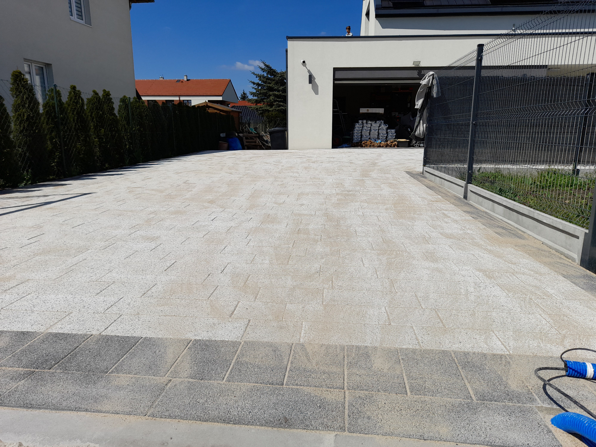 Nowo ułożony podjazd z jasnej kostki betonowej dekoracyjnej, obramowany ciemnoszarą kostką, prowadzący do garażu nowoczesnego domu z szarym ogrodzeniem.