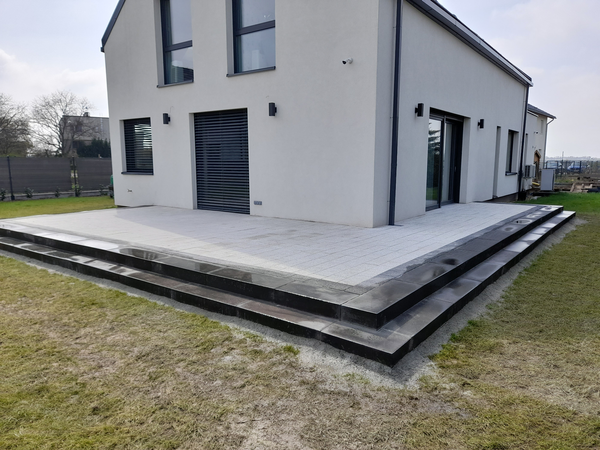 Nowoczesny taras z jasnoszarej kostki brukowej z czarnymi stopnicami schodowymi, przylegający do białego domu z szarymi oknami i roletami zewnętrznymi.