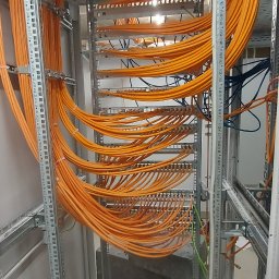 Energimixxxduo. S.c - Pomarańczowe kable ułożone w metalowej szafie rack, widoczne metalowe wsporniki, na dole porozrzucane kartony i plastikowe opakowania.