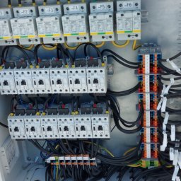 Energimixxxduo. S.c - Otwarta szafa elektryczna z zainstalowanymi bezpiecznikami, wyłącznikami i listwami zaciskowymi, widoczne liczne przewody w kolorze czarnym i żółto-zielonym, tło z widokiem na parking z pojazdami.