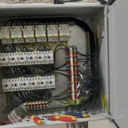 Otwarta skrzynka rozdzielcza z zainstalowanymi bezpiecznikami, stycznikami i listwami zaciskowymi, widoczne przewody oraz narzędzia: śrubokręty i szczypce.