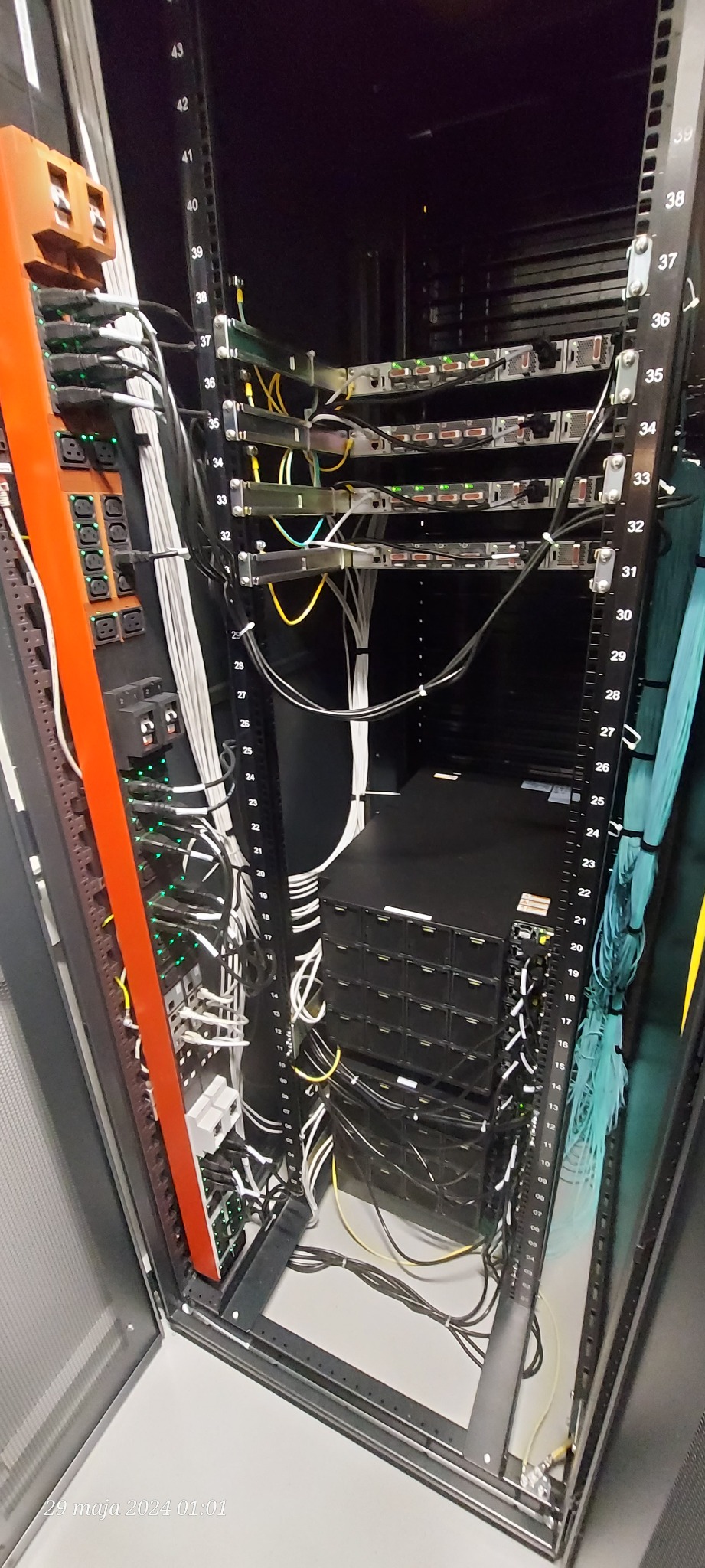 Serwerownia: szafa rack z panelami krosowniczymi, patch panele, okablowanie strukturalne w kolorach białym, żółtym i turkusowym, zasilacze awaryjne UPS, listwa zasilająca w kolorze pomarańczowym.