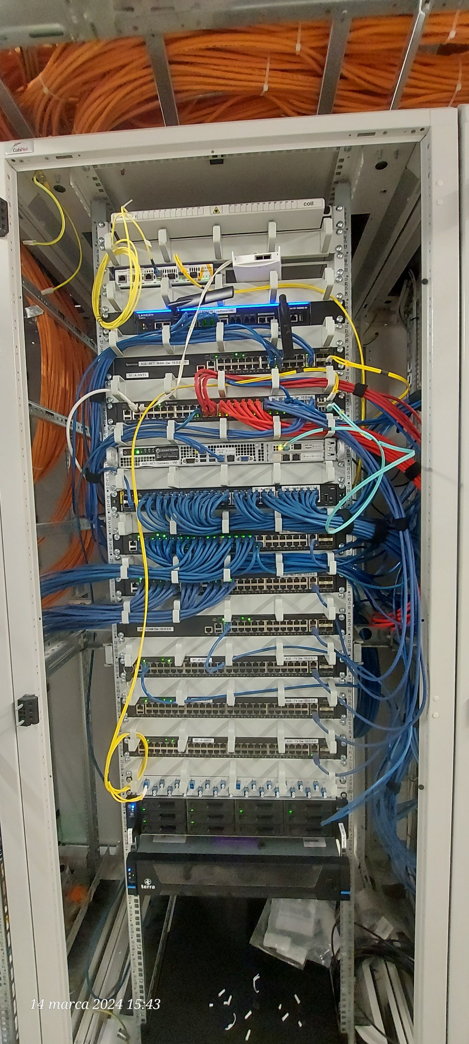 Szafa serwerowa z licznymi switchami i okablowaniem sieciowym w kolorach niebieskim, żółtym i czerwonym, widoczne pomarańczowe kable prowadzone górą, oświetlenie LED sygnalizuje pracę urządzeń.
