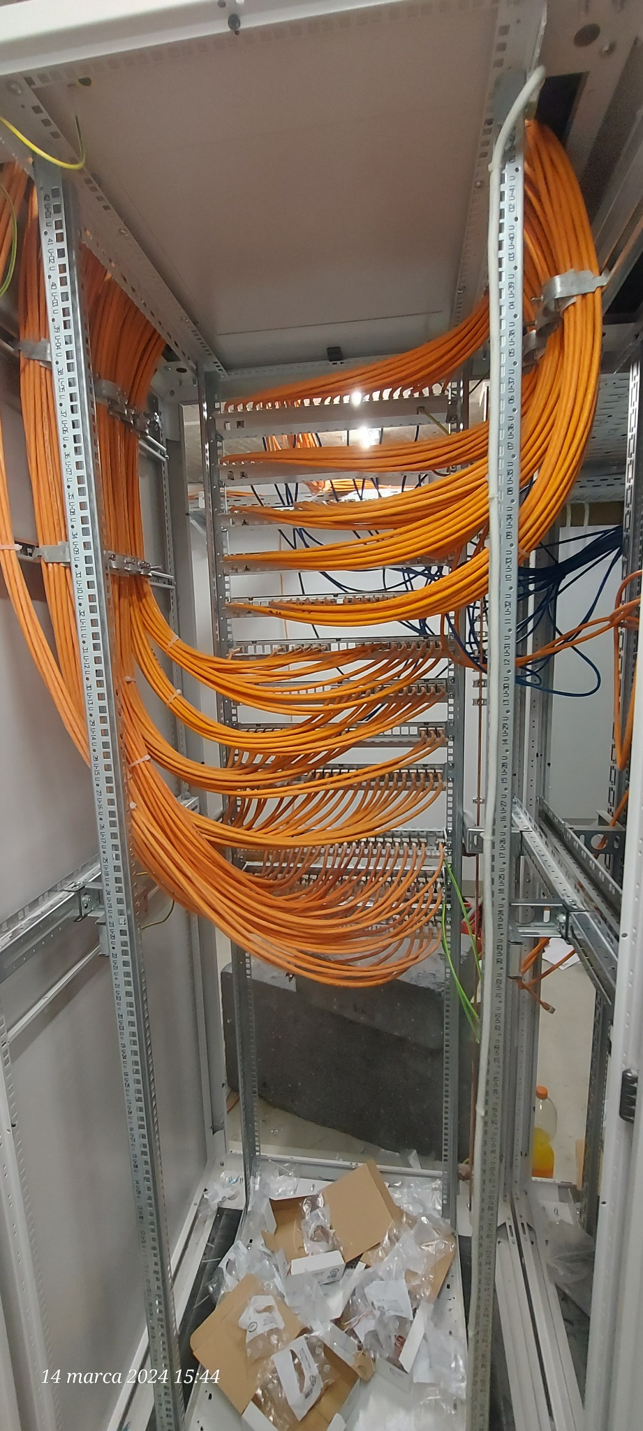 Pomarańczowe kable ułożone w metalowej szafie rack, widoczne metalowe wsporniki, na dole porozrzucane kartony i plastikowe opakowania.