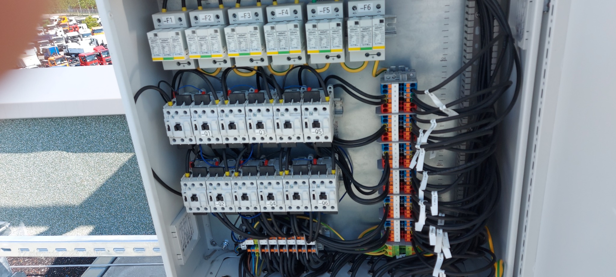 Otwarta szafa elektryczna z zainstalowanymi bezpiecznikami, wyłącznikami i listwami zaciskowymi, widoczne liczne przewody w kolorze czarnym i żółto-zielonym, tło z widokiem na parking z pojazdami.