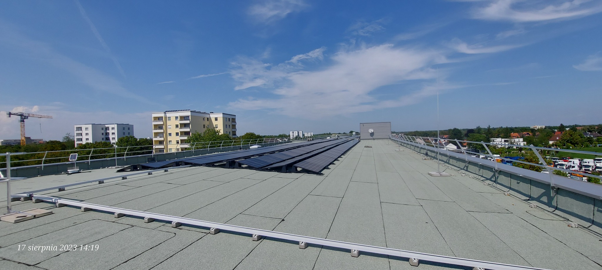 Widok z góry na płaski dach budynku z zainstalowanymi panelami fotowoltaicznymi i systemem odgromowym, w tle budynki mieszkalne i żuraw budowlany.