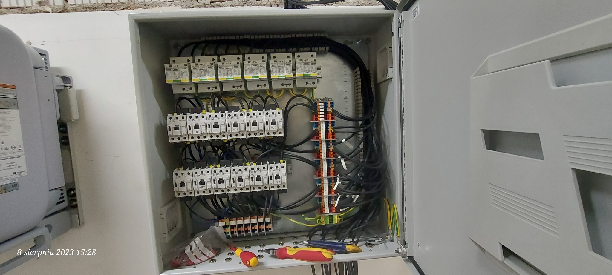 Otwarta skrzynka rozdzielcza z zainstalowanymi bezpiecznikami, stycznikami i listwami zaciskowymi, widoczne przewody oraz narzędzia: śrubokręty i szczypce.