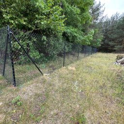 GARD Systemy Ogrodzeniowe Łukasz Zaręba - Czarna siatka ogrodzeniowa na metalowych słupkach, oddzielająca zarośnięty teren od lasu. W tle widoczne składowane drewno.