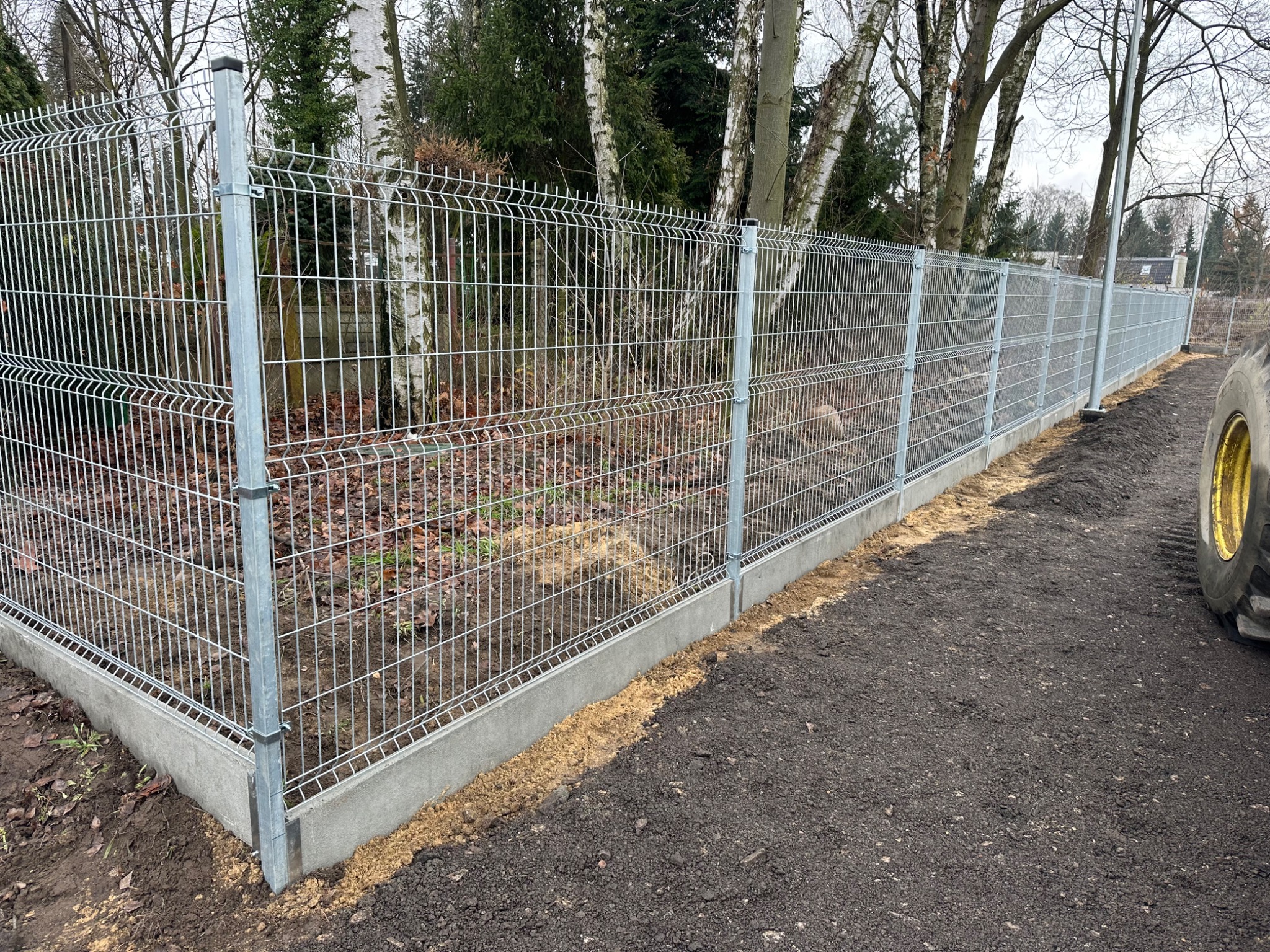 Nowoczesne ogrodzenie panelowe z podmurówką betonową, widok z boku, z fragmentem koła traktora po prawej stronie kadru. Tło stanowią drzewa.