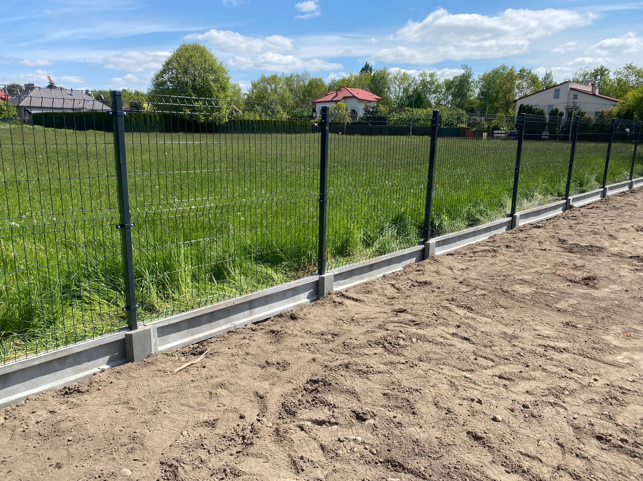 Nowoczesne ogrodzenie panelowe z podmurówką betonową na tle zielonego trawnika i zabudowy jednorodzinnej w słoneczny dzień. Widok z bliska na fragment ogrodzenia.